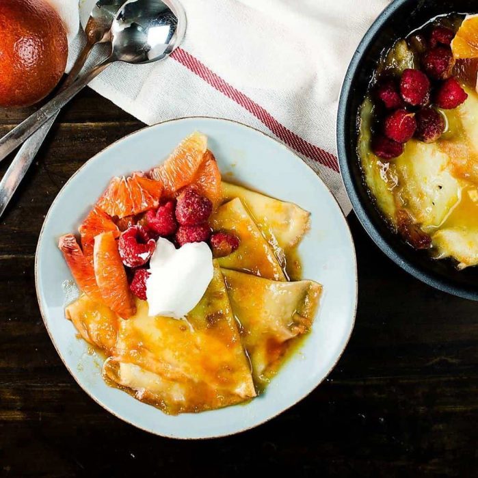 Blood orange crepes suzette on a plate, served with blood orange segments, raspberries, and whipped cream.