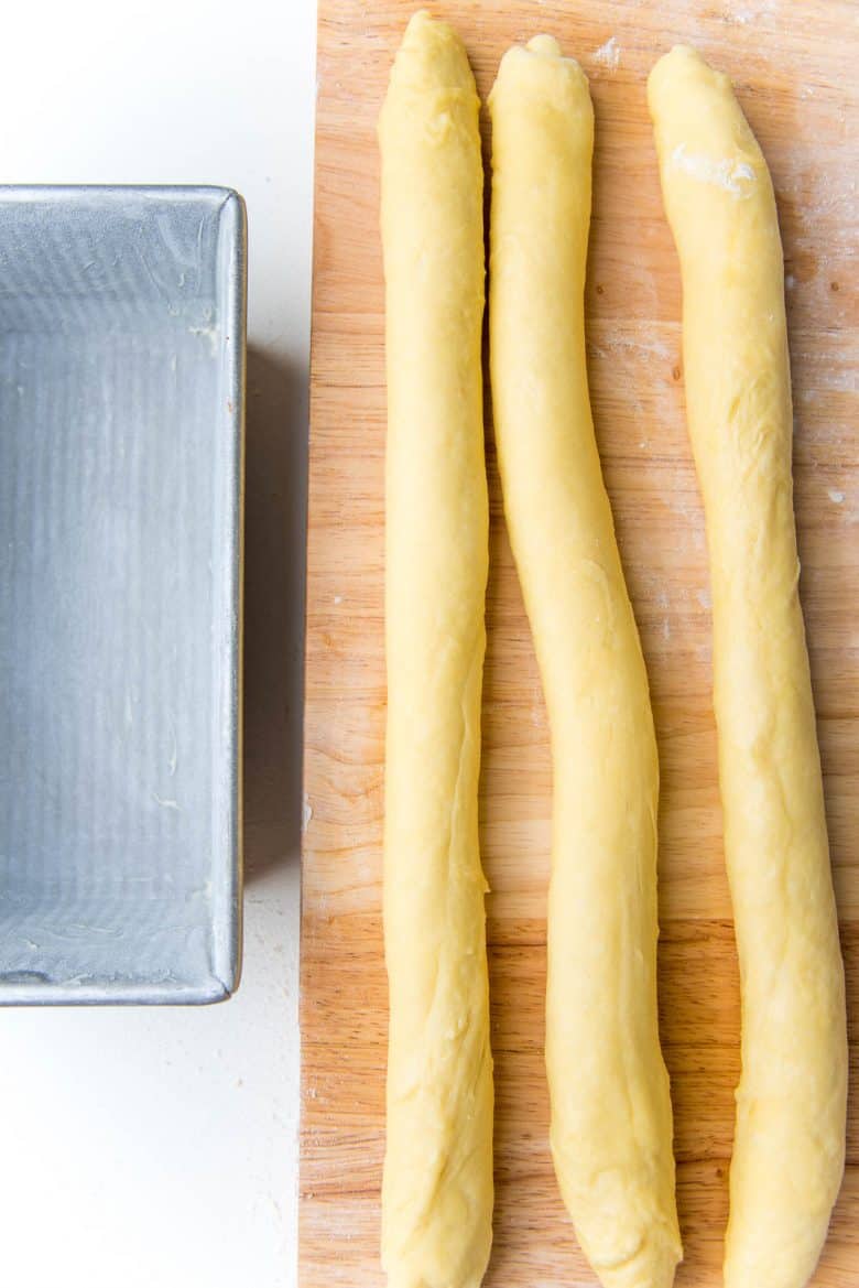 Three long ropes of dough on a wood work surface and a metal loaf pan on the side.