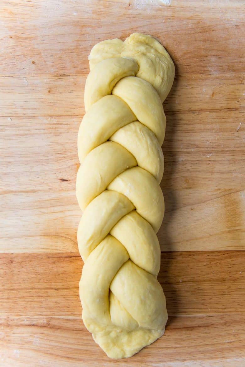 A braided brioche loaf dough on a wood work surface with the two ends flattened.