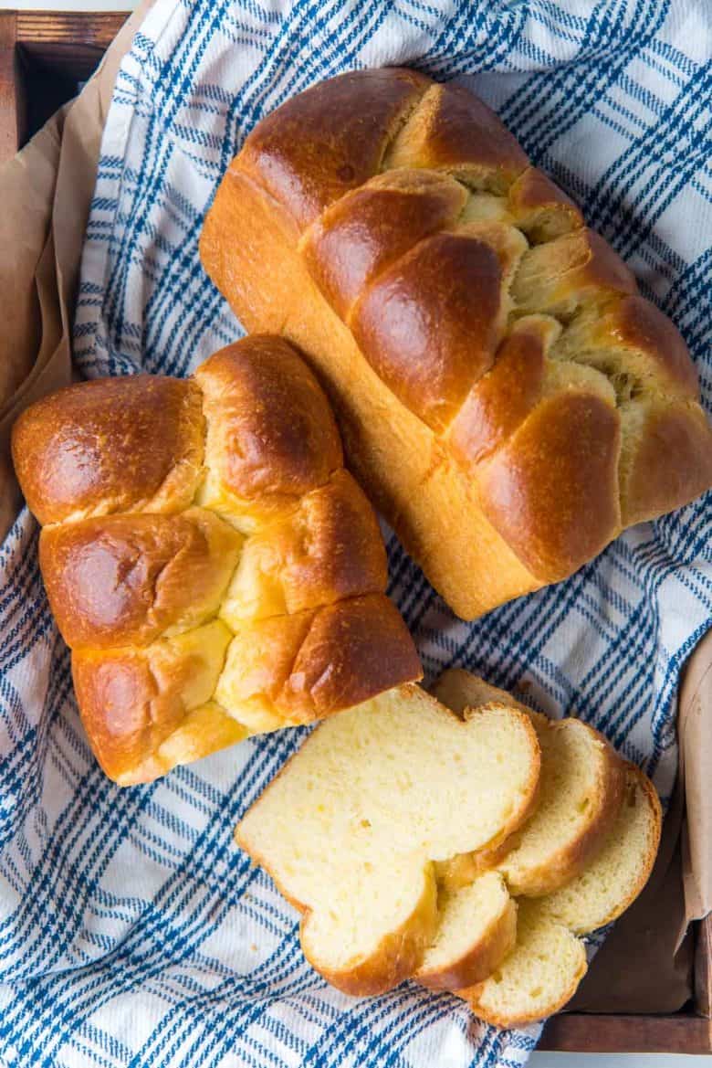 Two braided baked brioche bread loaves, where one loaf is sliced.