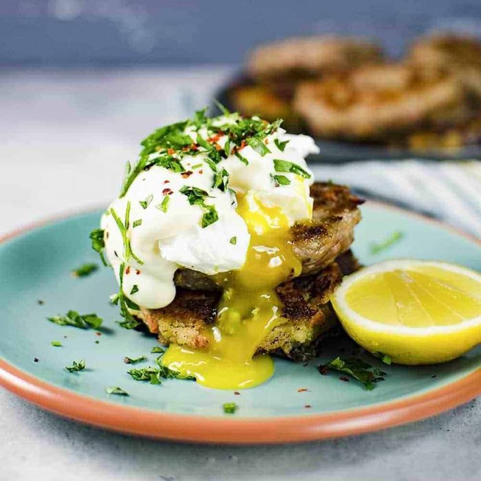 Two pan-fried bubble and squeak patties (potato cakes) on a small plate with a poached egg and herbs on top.