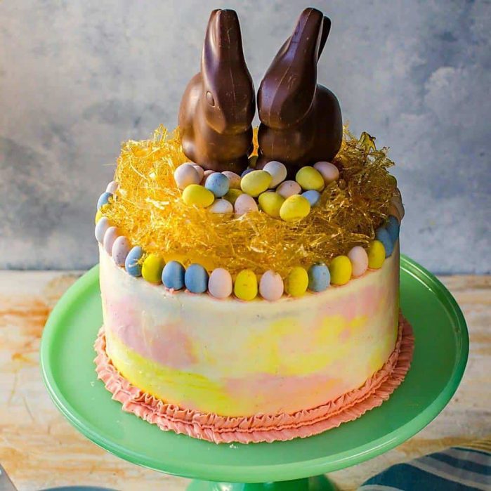 Colorful vanilla chai cake with Easter decorations and chocolate bunnies on top, on a cake stand.