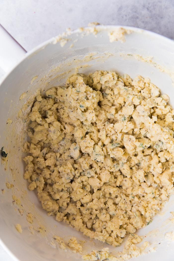Recipe process for making thyme and cheddar cheese cookies - Crumbles of the cheese cookie after the flour has been mixed through in a white bowl.
