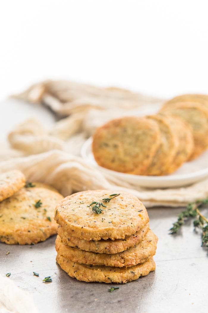 A stack of thyme and cheddar cheese cookies placed in the foreground with more cheddar cookies in the background on a white platter, with sprigs of thyme. 
