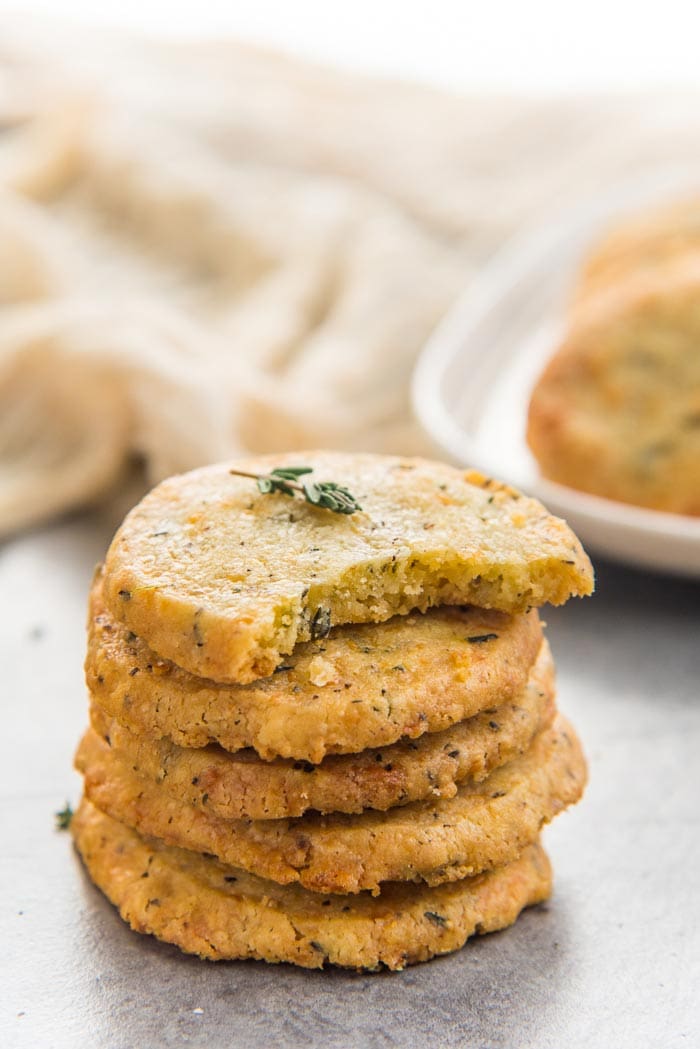A stack of thyme and cheddar cheese cookies with 4 whole cookies and a bite taken out of the top cookie, with more cookies in the background. 