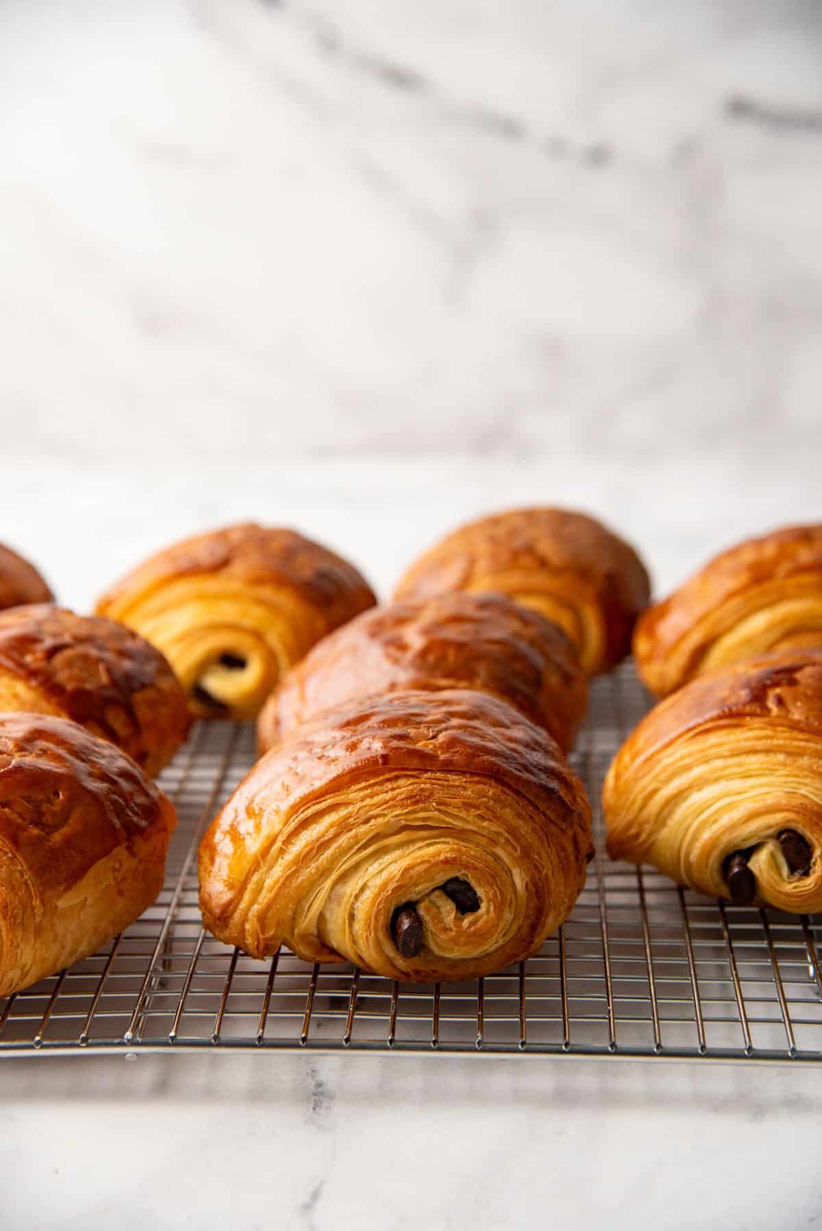 Several baked, golden brown, flaky chocolate croissants on a metal wire rack.
