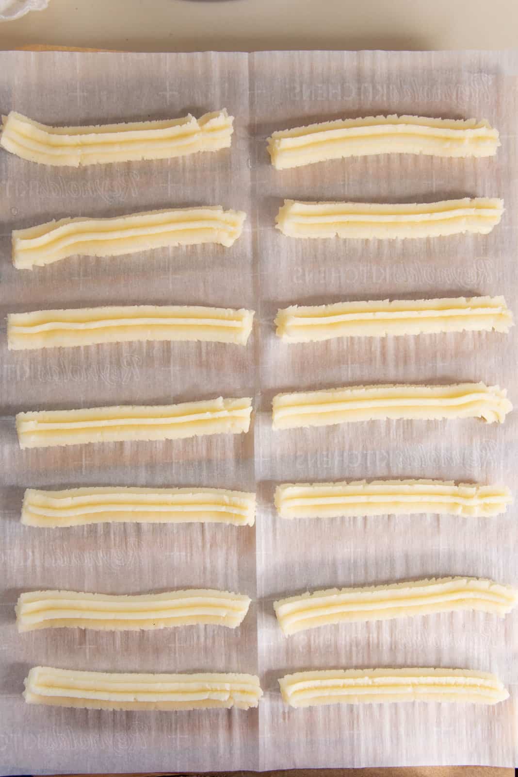 14 pieces of raw churro dough piped out in lines on a parchment paper.