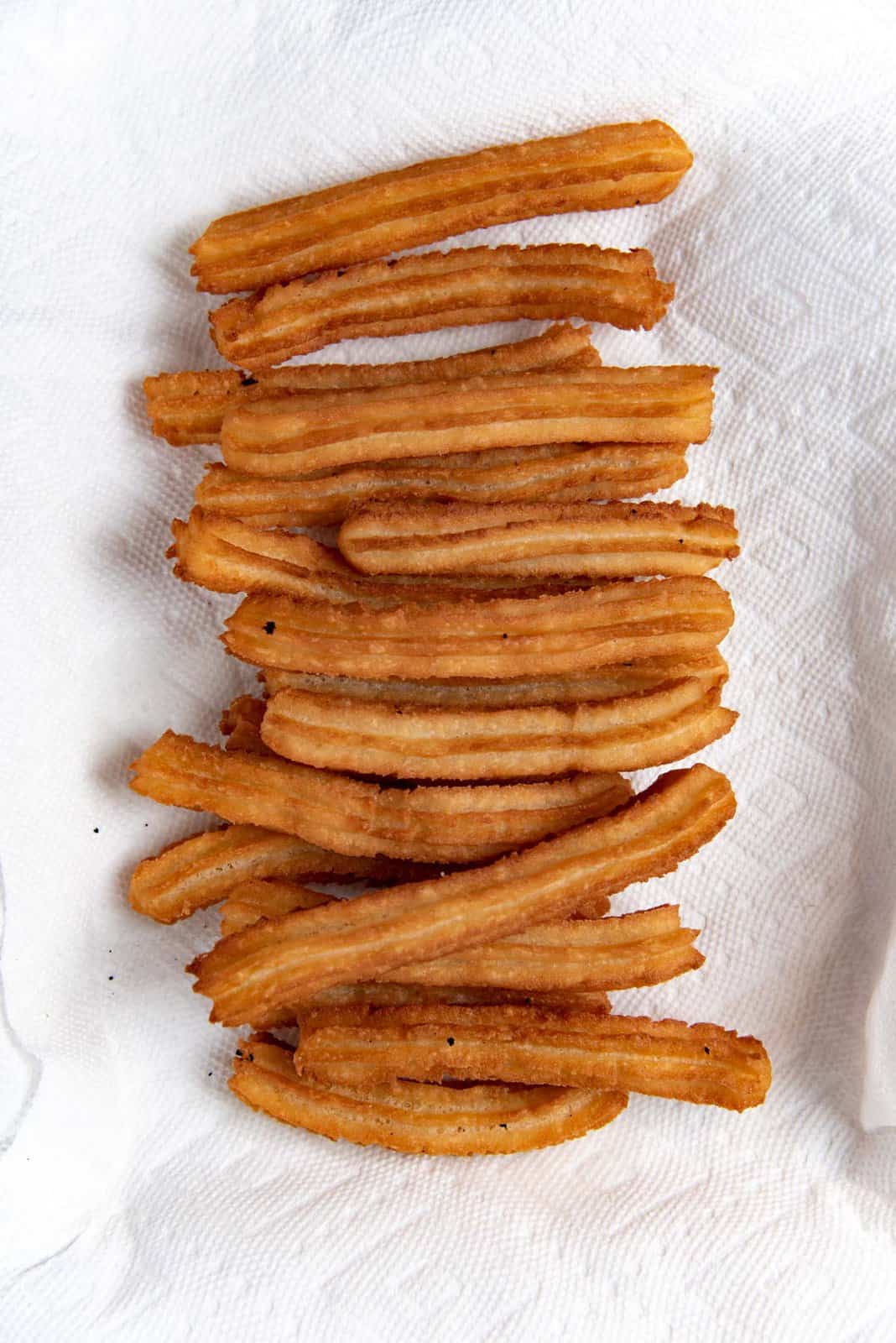 Overhead image of 5 to 6 inch long churro sticks fried to a golden brown color and stacked on a paper towel to drain.