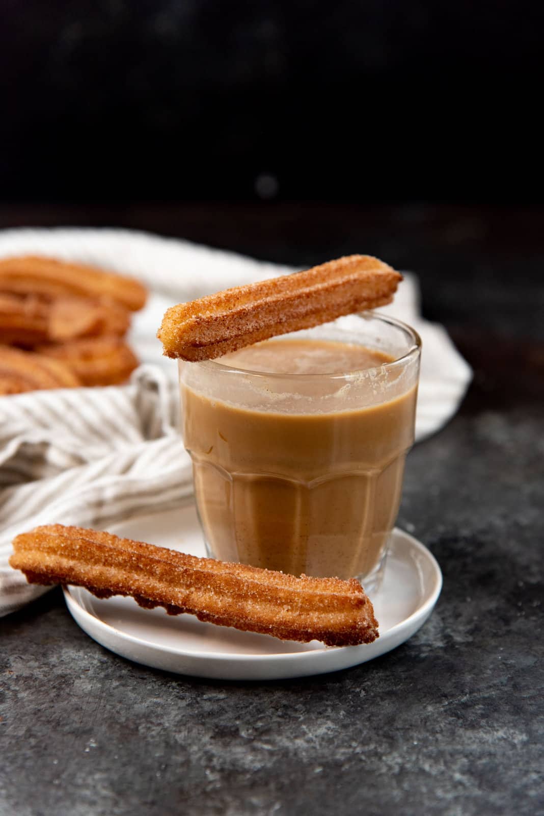 A coffee in a small glass, served on a small white saucer, with one churro placed ontop of the glass rim, and another churro placed on the saucer next to the glass.