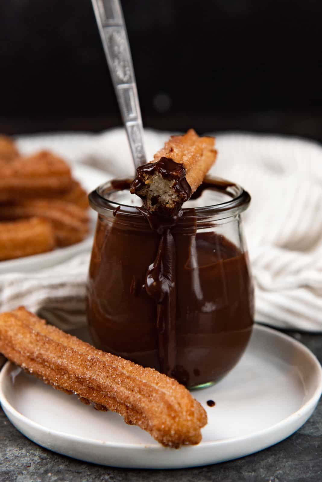 A close up of a small pot of melted chocolate sauce with a half eaten churro on top, and an uneaten churro placed next to the chocolate pot.