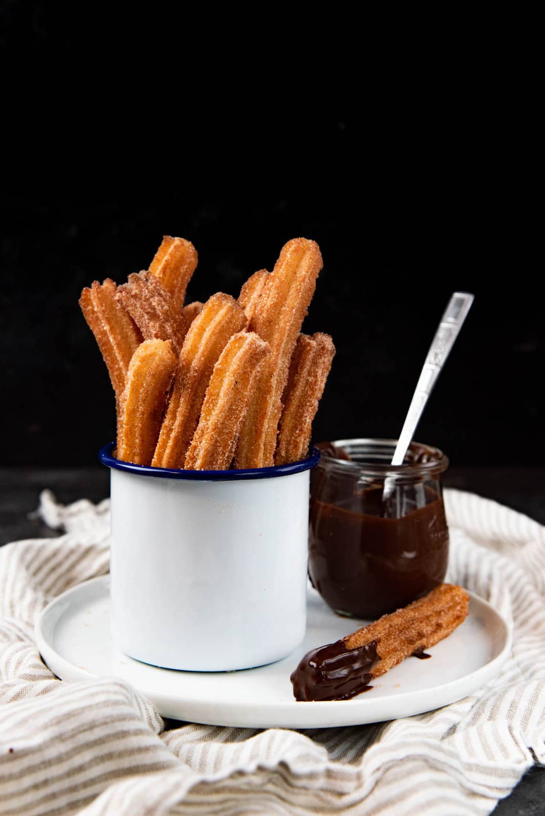 Authentic churros stacked inside a white mug, standing up like fries, served on a white plate with a chocolate pot and a chocolate dipped churro next to the white mug.