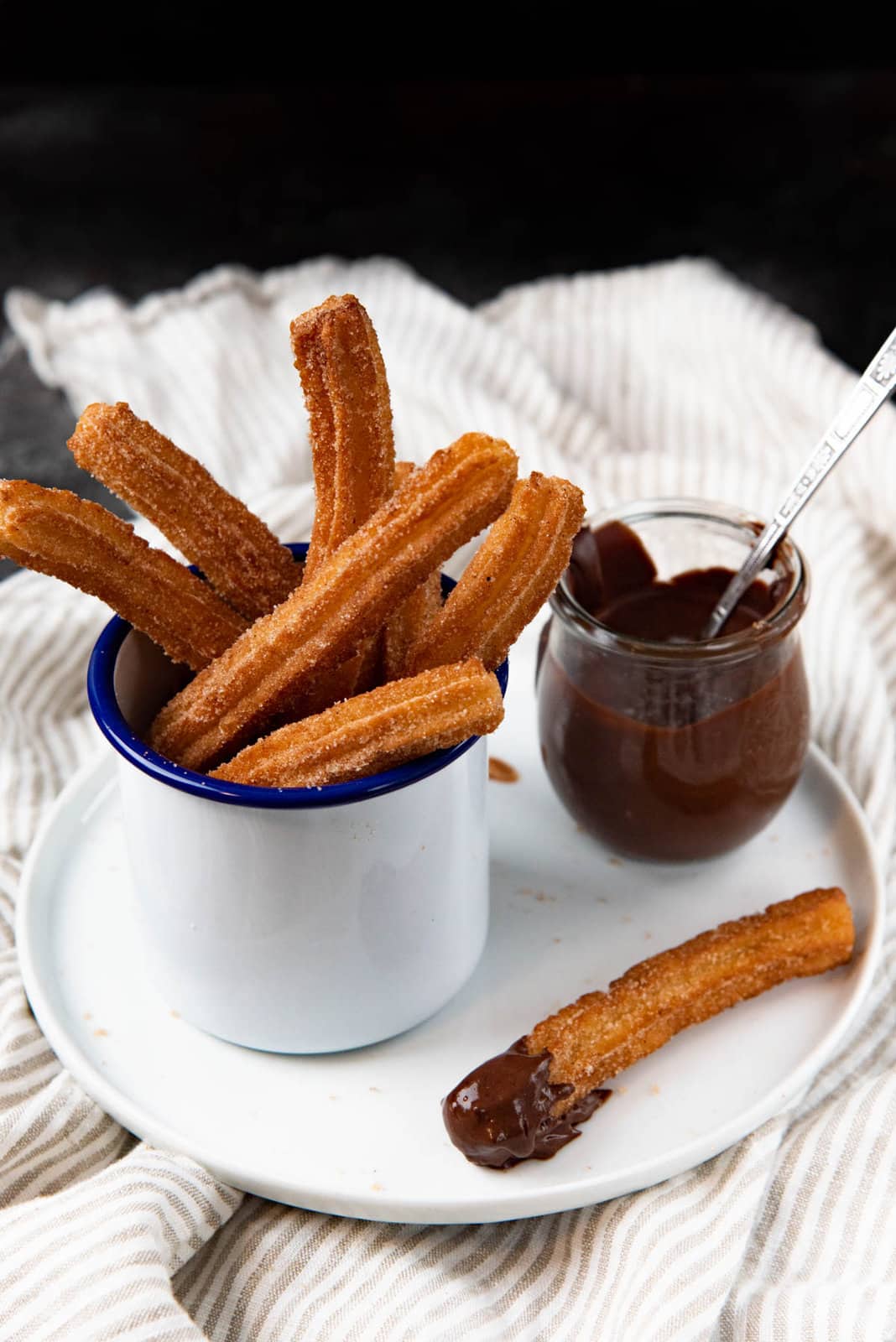 An angled view of churros served in a white enamel mug, and a chocolate sauce pot, and a dipped churro next to it, all on a white plate.