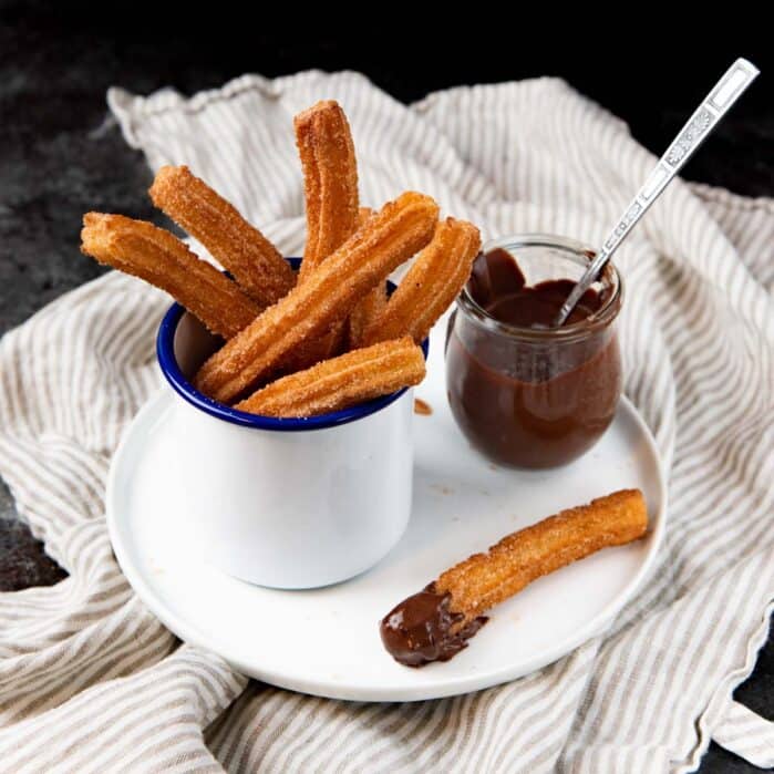 Square image of An angled view of churros served in a white enamel mug, and a chocolate sauce pot, and a dipped churro next to it, all on a white plate.