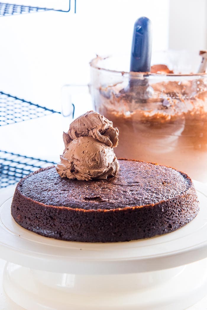 One Chocolate cake layer on a cake decorating stand, with scoops of chocolate butter cream on top. The rest of the chocolate frosting in a bowl in the background.