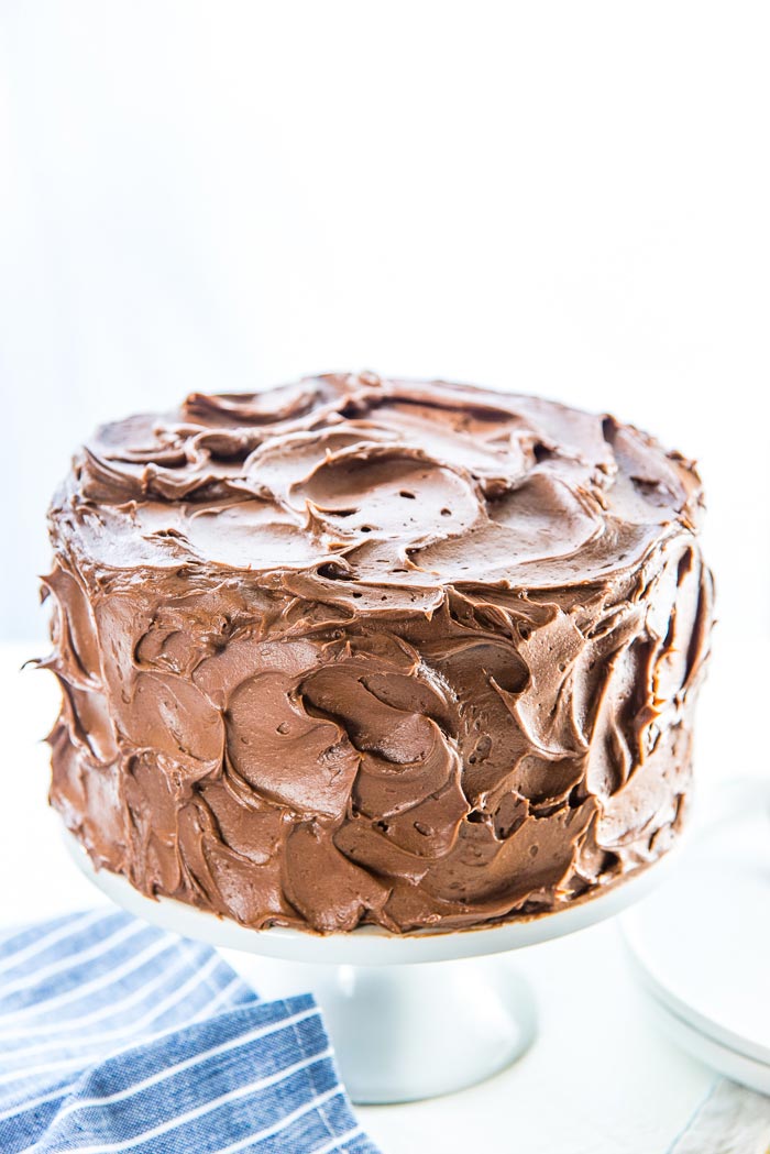 A completely frosted classic chocolate cake, on a white cake stand. 