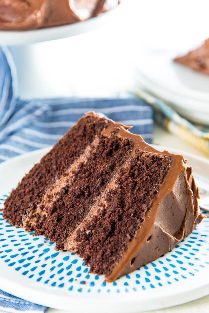A close up of the slice of classic chocolate cake, lying on the side, on a blue spotted plate. With 3 layers of cake and a creamy chocolate frosting. 