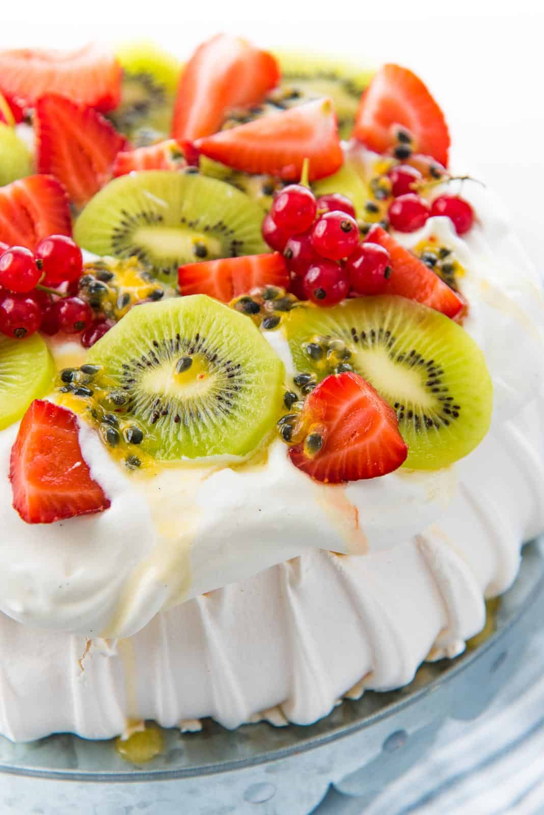 A close up of a pavlova on a cake stand, topped with whipped cream, kiwi fruit, strawberries, passion fruit and redcurrants