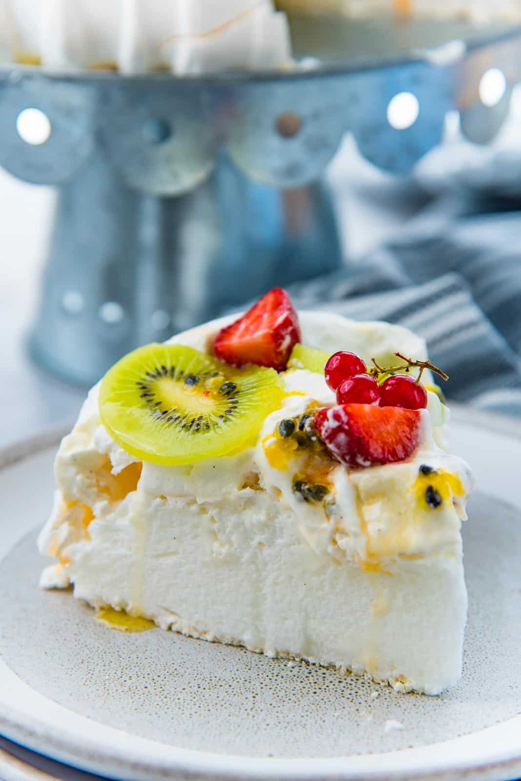 A close up of a pavlova slice topped with whipped cream and fruits, on a grey plate.