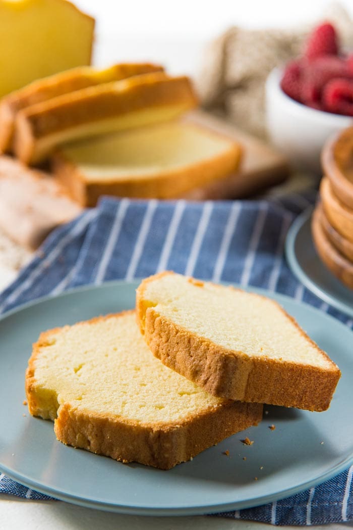 two slices of classic pound cake on a blue plate, with the rest of the pound cake, sliced, in the background. with a bowl of berries and more serving plates in the background. 