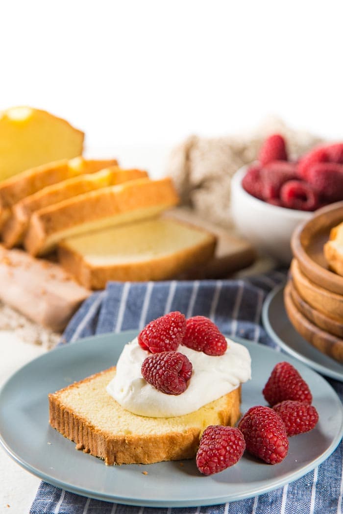 A slice of pound cake on a blue plate, served with a dollop of whipped cream on top, with fresh raspberries. A bowl of fresh raspberries, more sliced pound cake in the background.