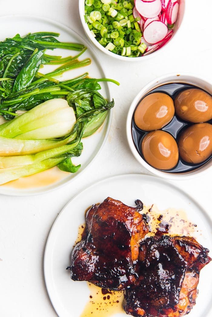 An assortment of simple ramen toppings on white plates - caramelized soy chicken, Sauteed Asian greens, sliced green onions, crunchy radishes, and ramen eggs.
