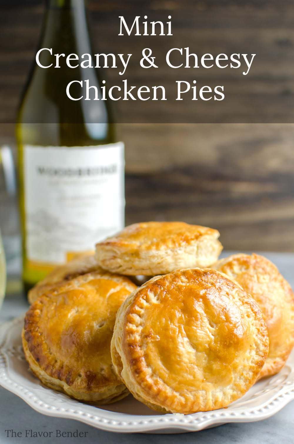 Several buttery, flaky mini puff pastry chicken pies on a white platter with a bottle of wine in the background. 