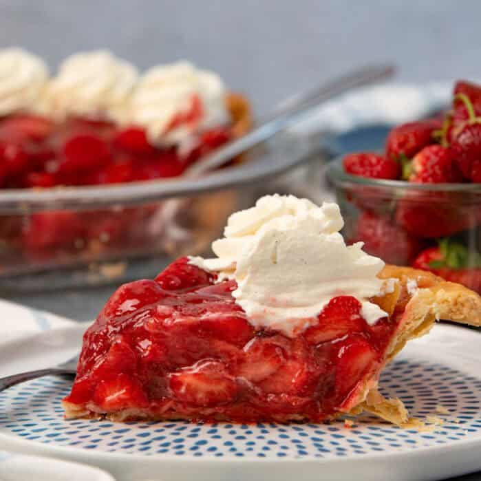 Fresh strawberry pie slice on a plate with whipped cream and the rest of the pie in the background.