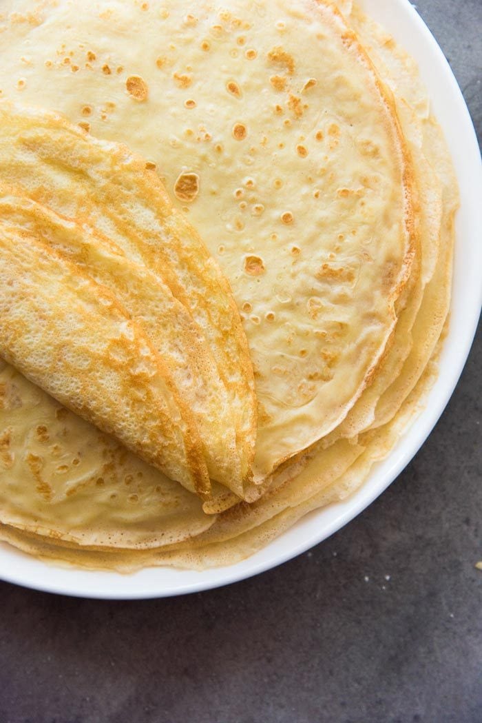A stack of cooked classic french crepes on a white plate, showing both caramelized sides of the crepes. 