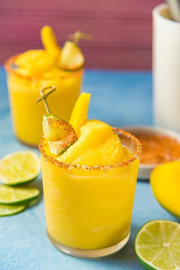 Frozen Mango Margarita served in a straight glass, with fresh mango garnish and a tajin seasoning dipped lime slice garnish. With lime slices, Fresh mango and tajin seasoning places on the blue table top.