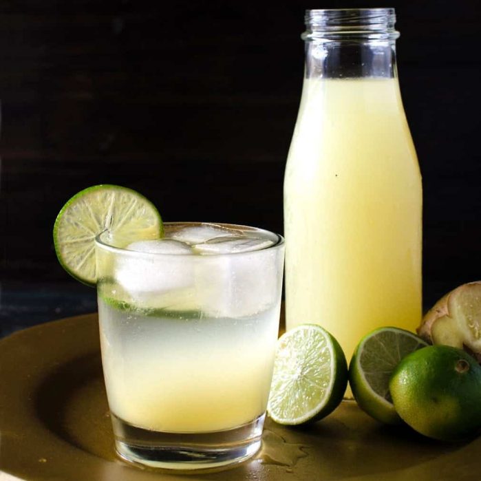 Homemade ginger limeade in a glass with several limes and a bottle of limeade in the background.