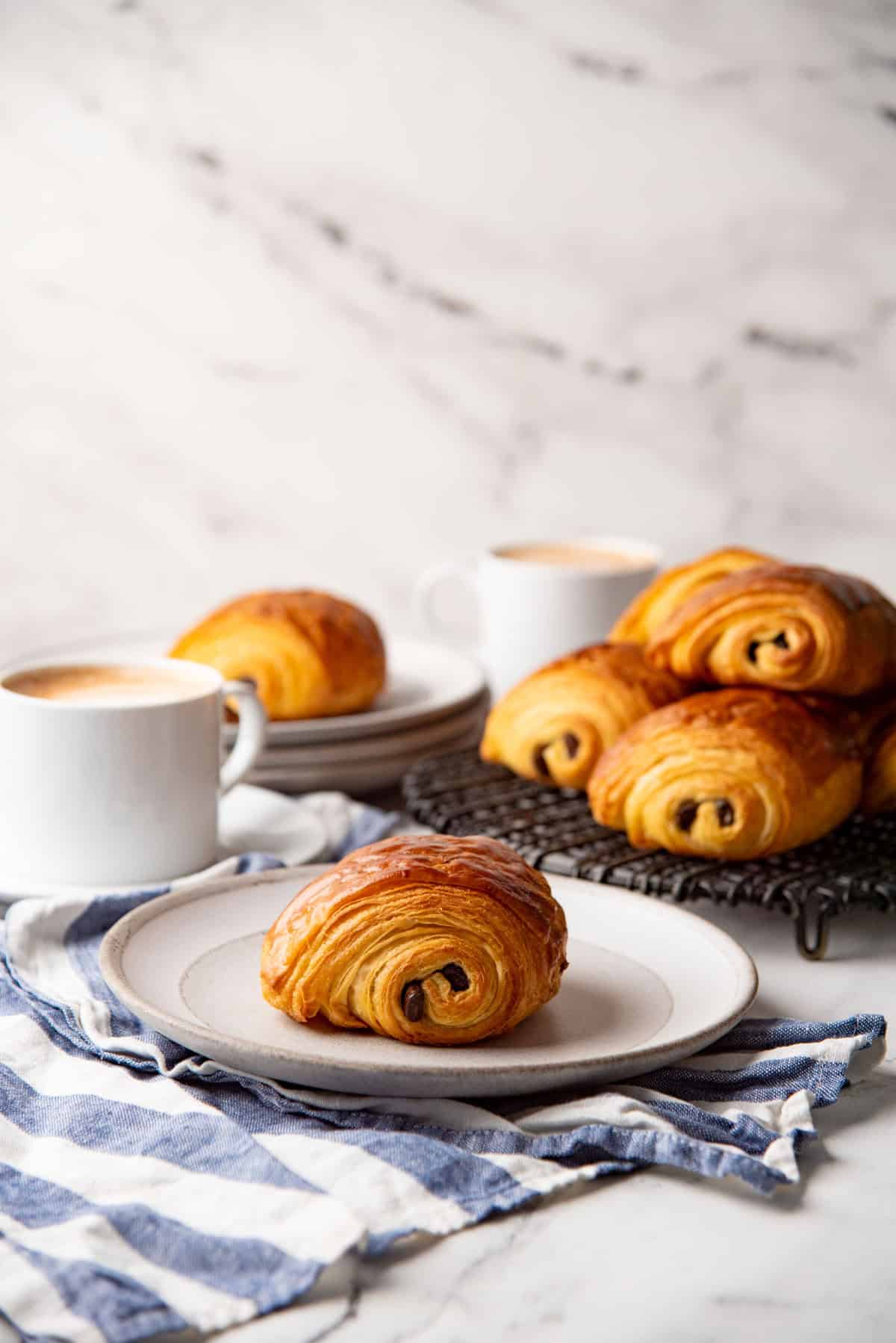 A chocolate croissant on a white plate, and more croissants on a wire rack in the background.
