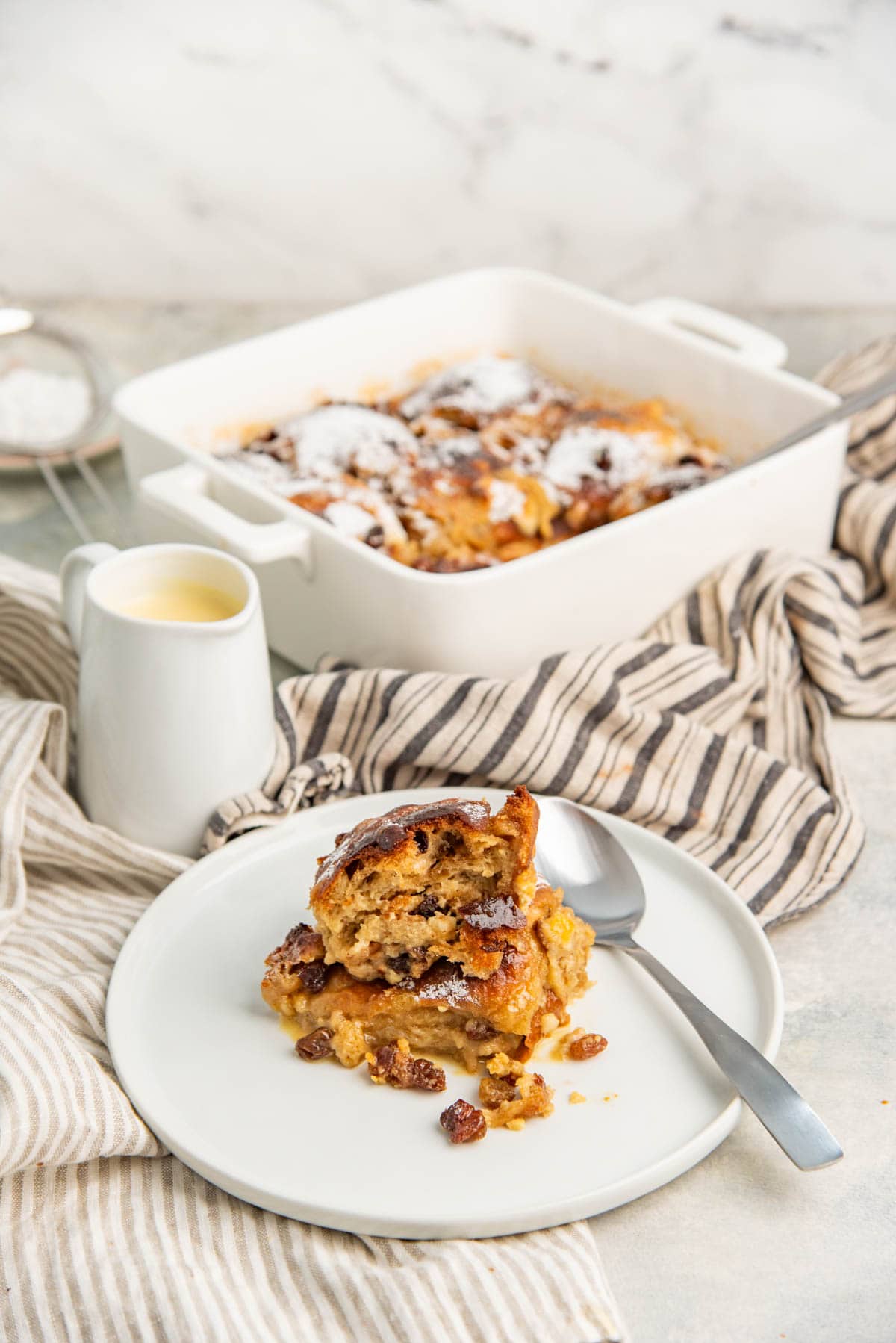 Baked hot cross bun bread pudding served in a small white plate with a small jug of pouring custard next to it.