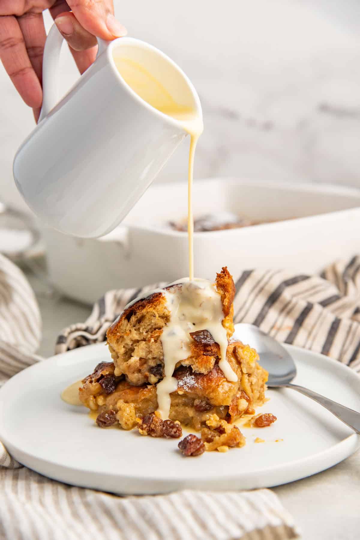 Pouring crème anglaise over baked hot cross bun pudding served in a small white plate with a spoon.