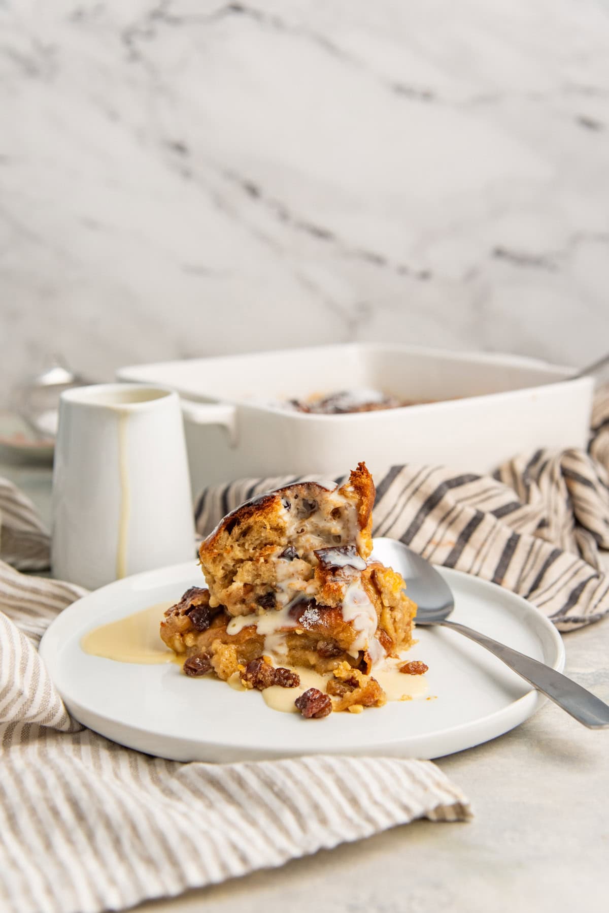 A serving of hot cross bun pudding on a white plate with vanilla crème anglaise and a spoon.
