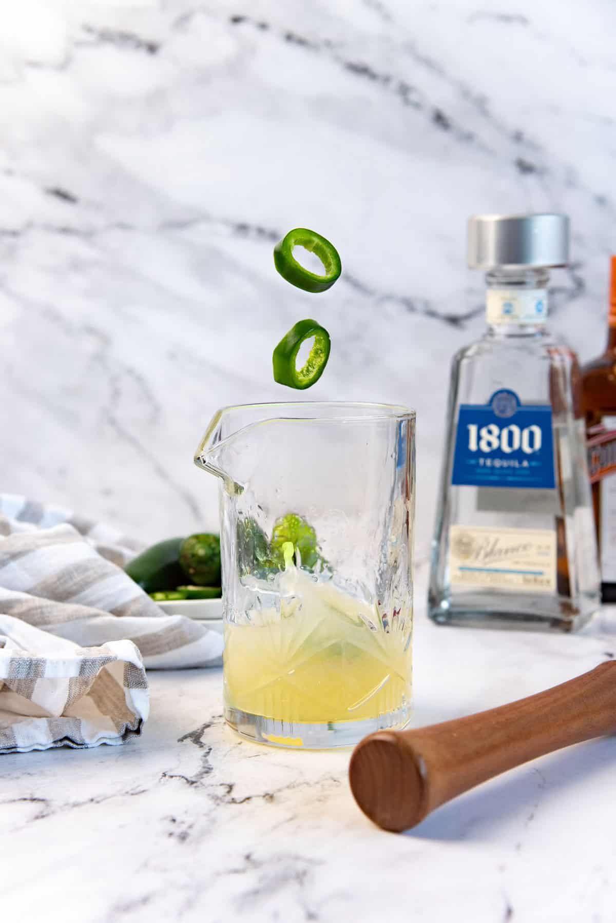 An image of two jalapeno slices falling into a cocktail mixing glass, with the liquid splashing, with bottles of tequila in the background.