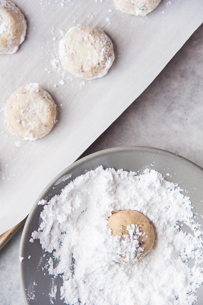 Freshly baked warm Mexican Wedding Cookies being coated with confectioners sugar in a bowl, and coated Mexican Wedding cookies on a baking tray