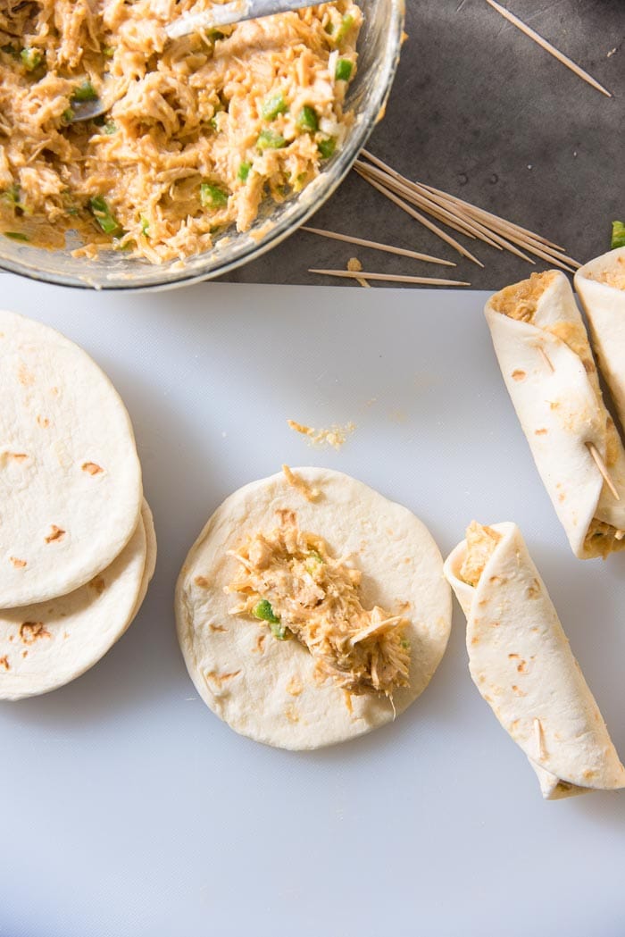 Mini tortillas being filled with the creamy chicken filling, with rolled taquitos in the background.