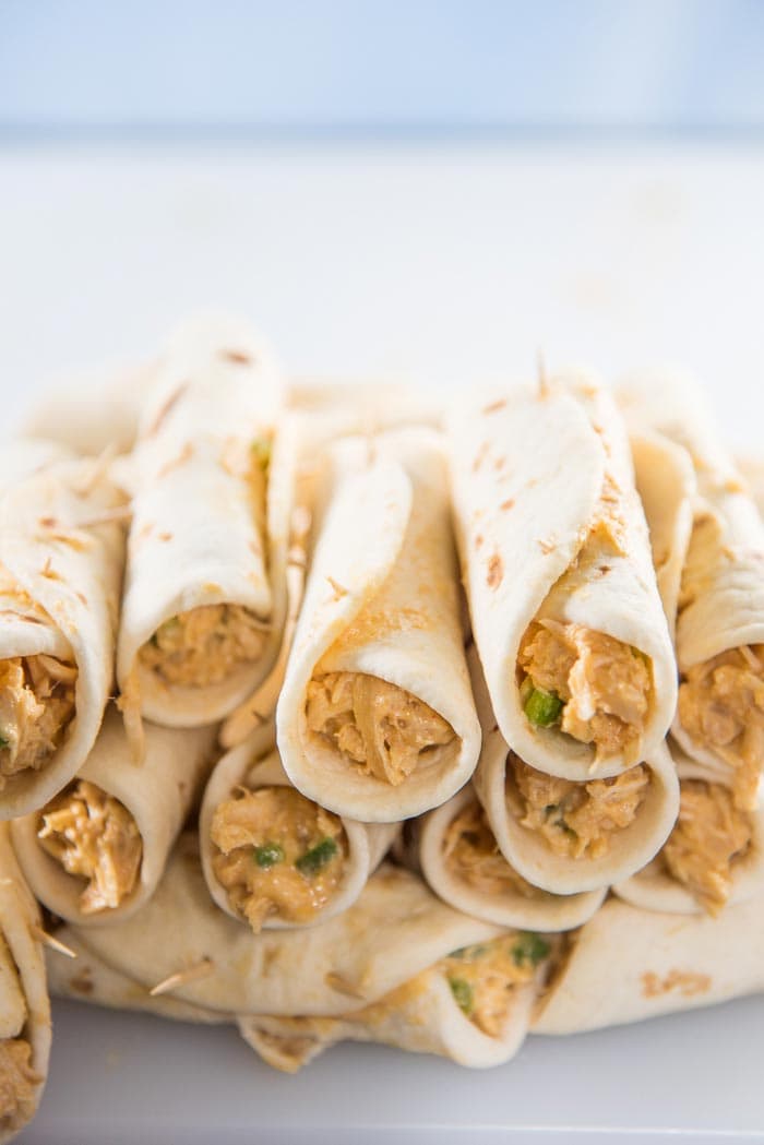 Rolled up mini taquitos, secured with toothpicks. Un fried taquitos stacked on top of each other before being fried.