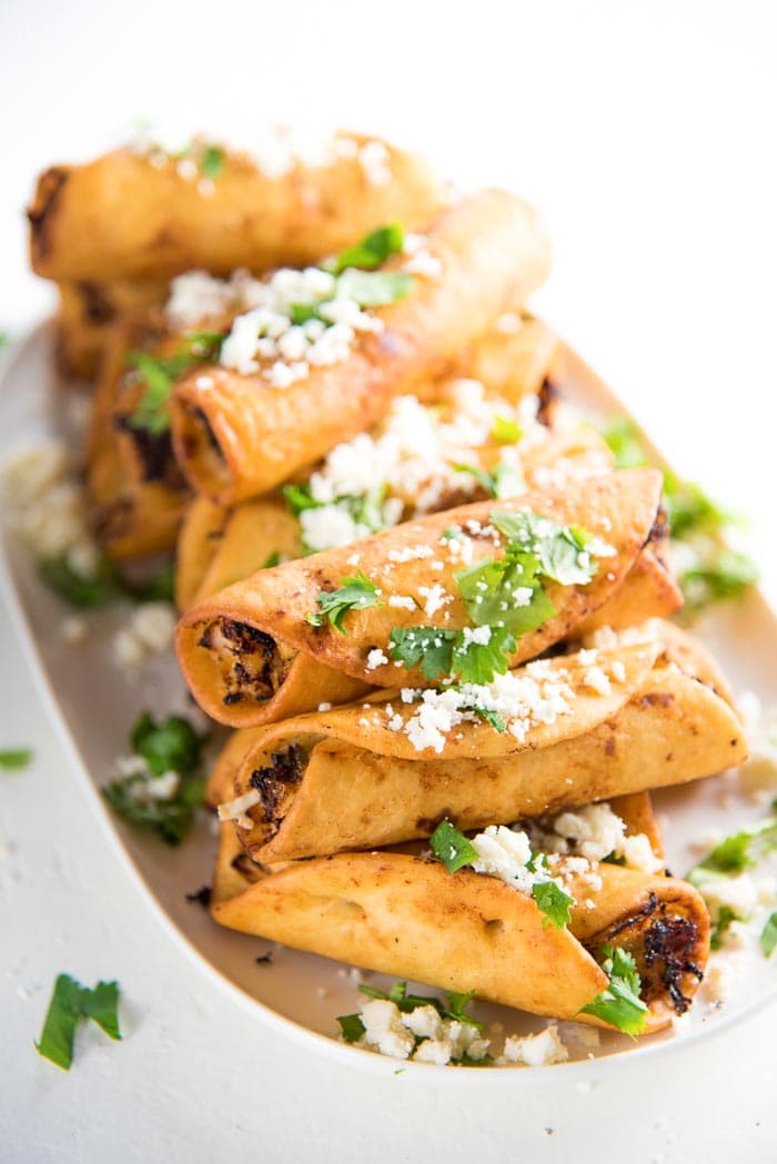 Crispy mini chicken taquitos placed on a white platter, with chopped cilantro and crumbled cojita on top