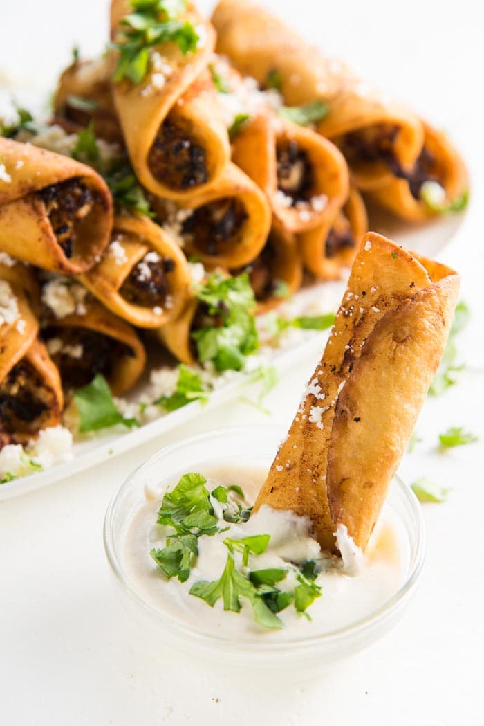 Mini Chicken taquito dipped in jalapeno sour cream, with chicken taquitos in the background on a platter, with cheese and cilantro sprinkled on top. 