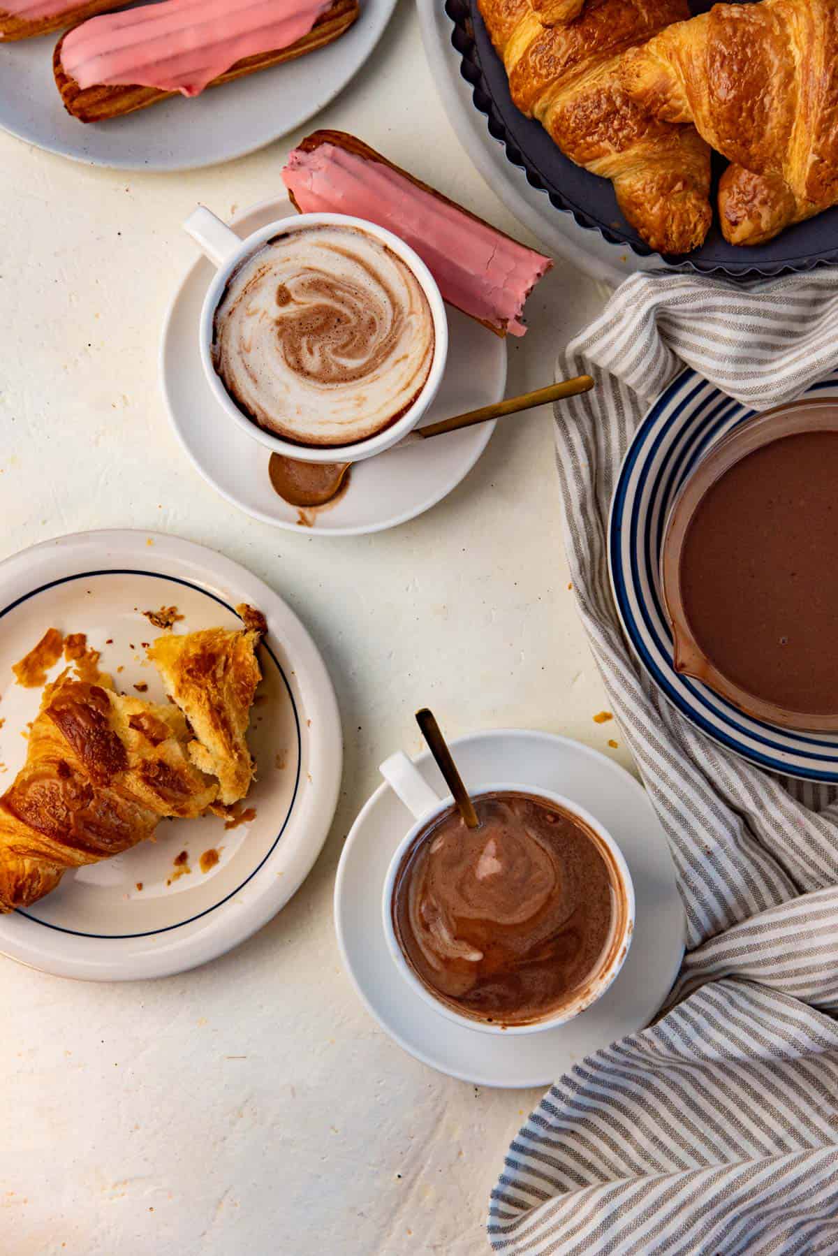 Overhead view of two cups of French hot chocolate served with homemade croissants and eclairs.