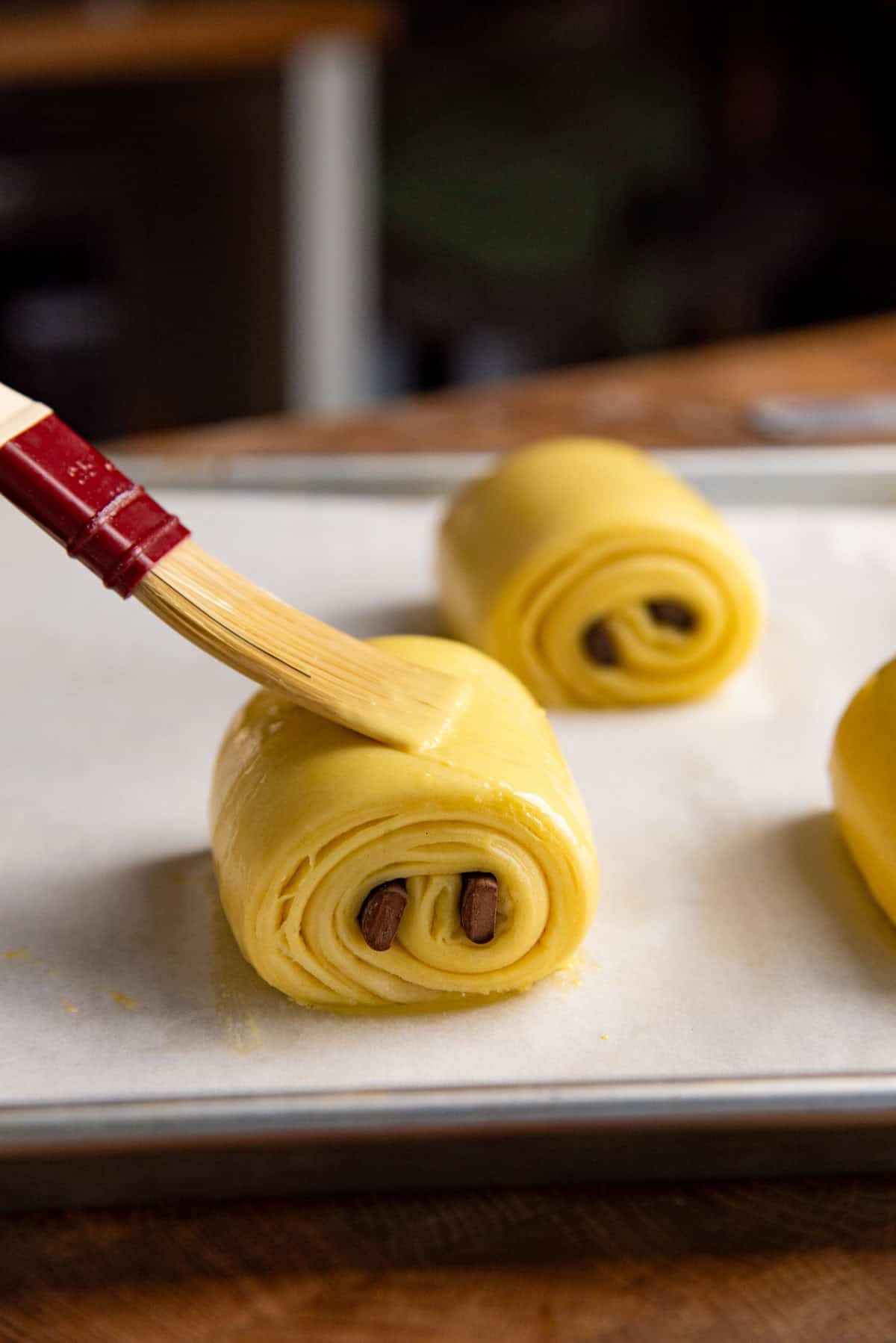 Brushing proofed chocolat croissants on a baking sheet with egg wash just before baking them.