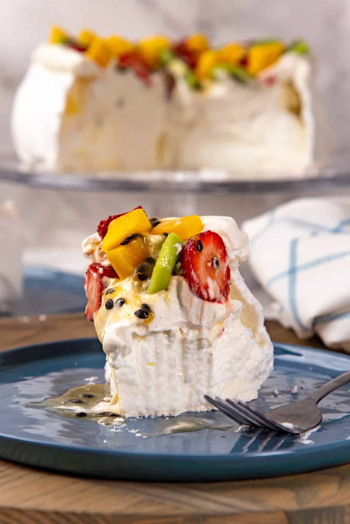A close up of a slice of pavlova served on a blue plate, with whipped cream and fresh fruit on top, with a piece taken to show the soft marshmallow center of the pavlova.