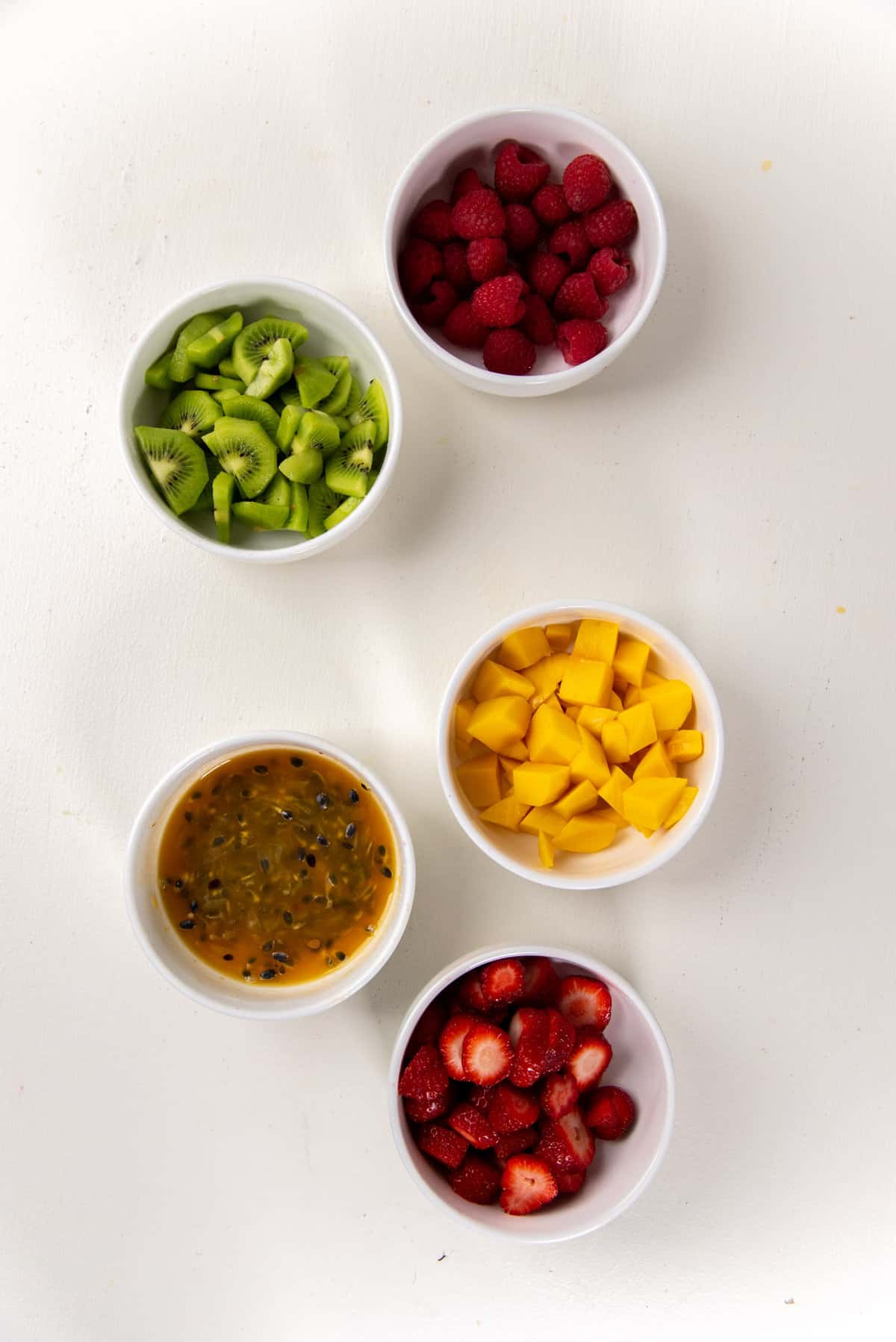 Fresh fruits peeled and prepared to use as topping for the pavlova, in separate bowls. From the top, raspberries, sliced and halved kiwi fruit, dived mangoes, passionfruit pulp in syrup, and sliced strawberries.