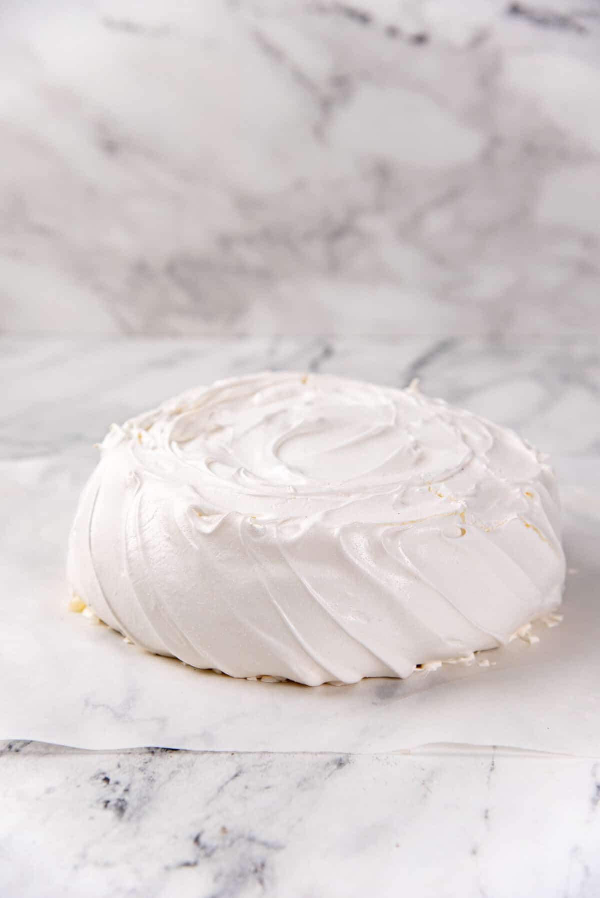 An image of a pavlova on a parchment paper, that has been cooled down placed on a marbled counter top.