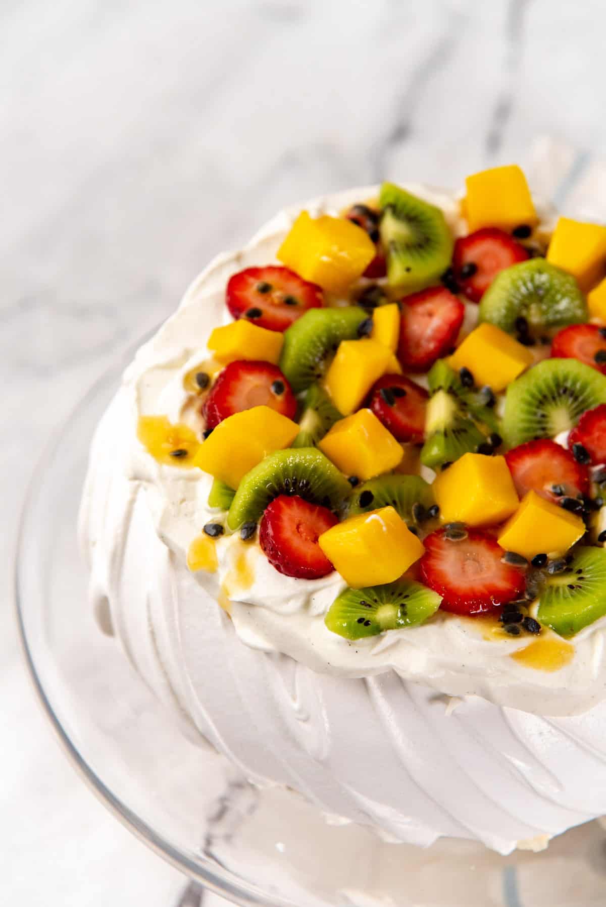 An angled overhead view of the pavlova topped with whipped cream and colorful fresh fruits piled on top.