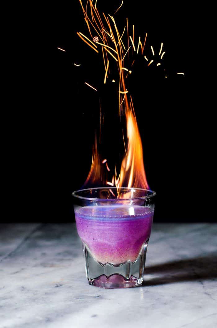 Cocktail glass with a purple drink with a blue layer on top and fire and sparks on the drink surface.