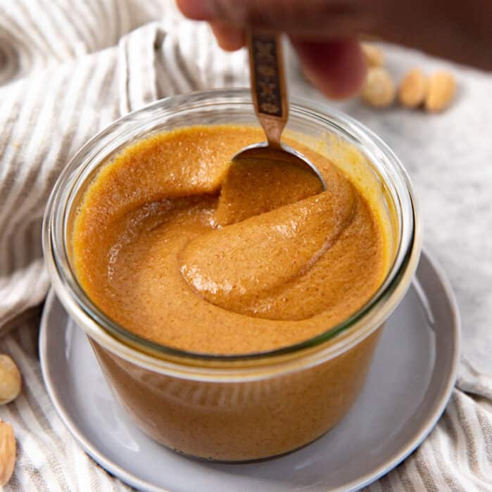 Square image of praline paste in a glass jar, being stirred with a spoon.