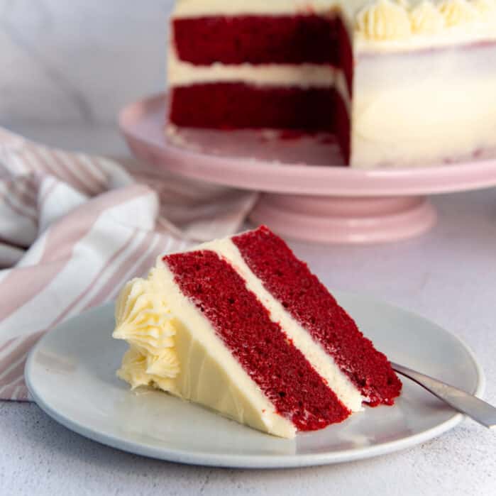 Slice of red velvet cake with cream cheese frosting on a light grey plate.