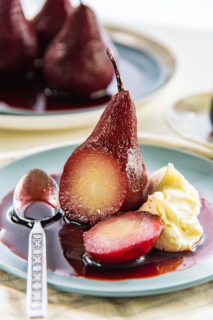 A single red wine poached pear on a blue plate with red wine syrup, with a piece cut off to show the middle, with a spoon on the side.